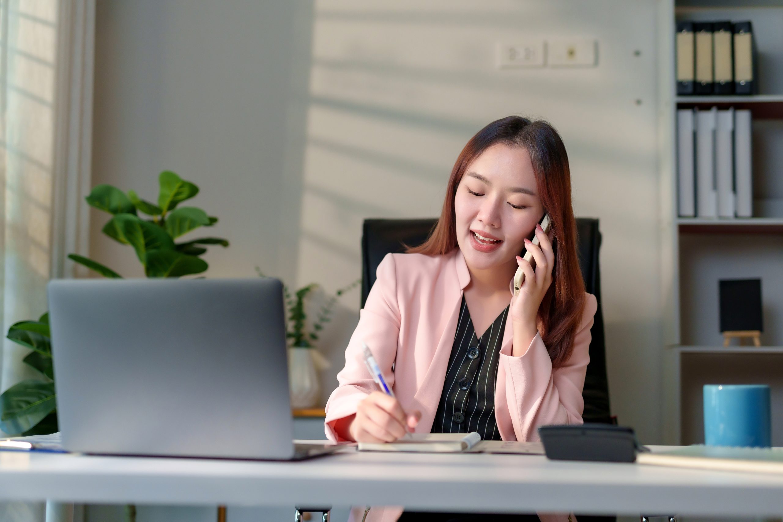 Young Asian businesswoman working in office, talking on mobile phone while writing in a notebook, managing multiple tasks